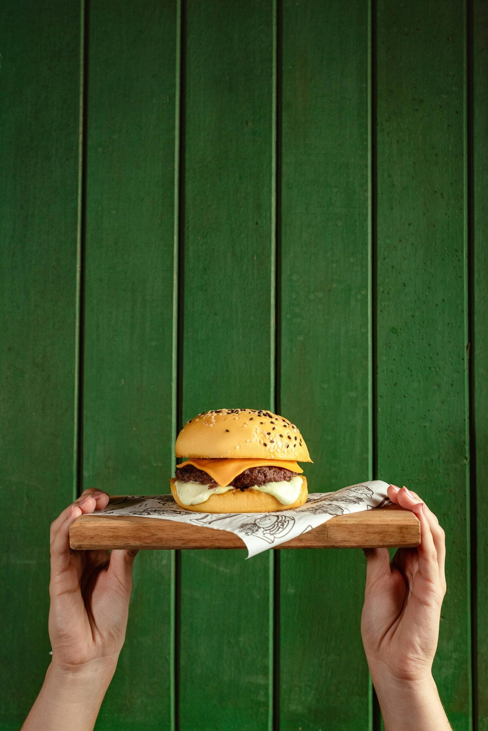 Delicious cheeseburger served on a wooden tray held by hands against a vibrant green background.
