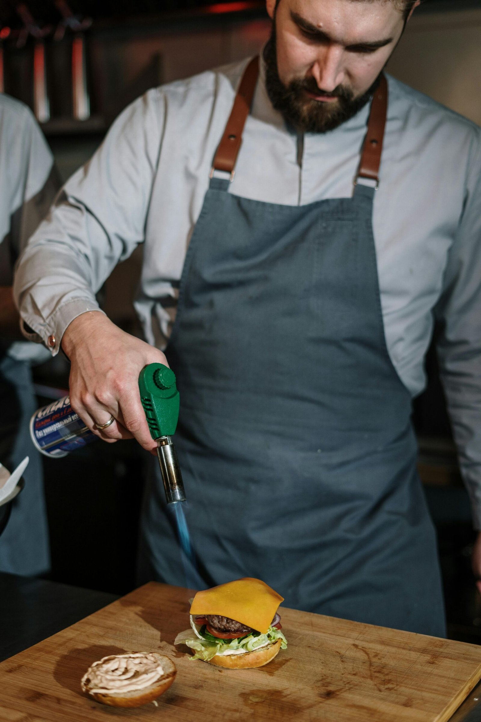 Chef uses blowtorch to melt cheese on gourmet burger in kitchen.